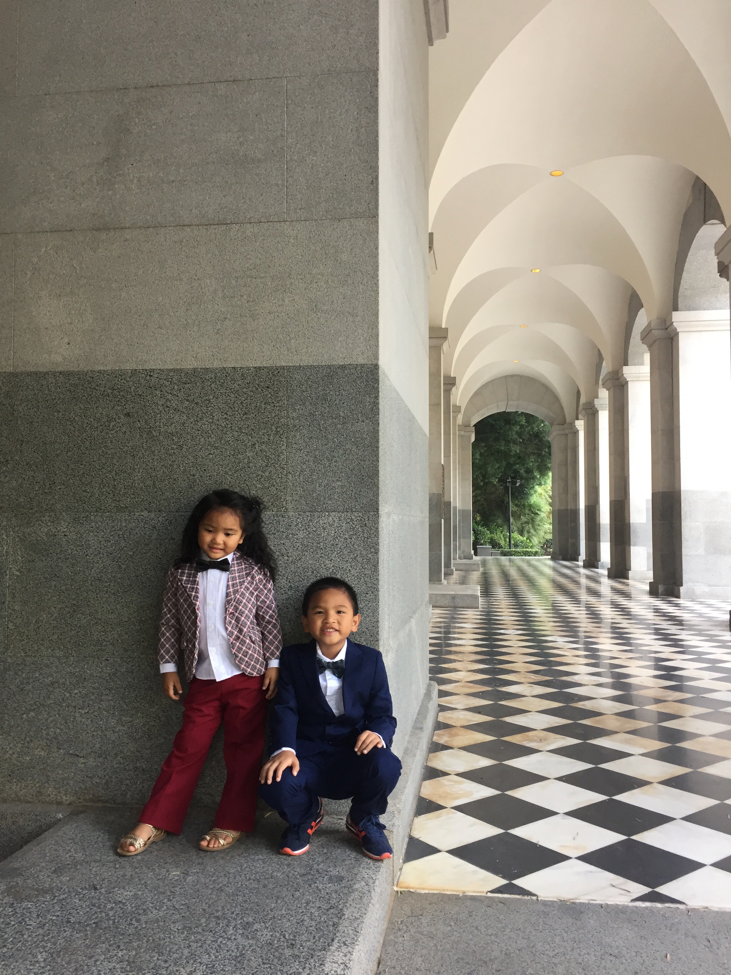 Two children dressed in formal attire pose beside a column in a covered arcade.