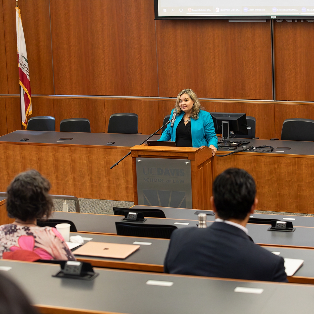 Professor Raquel Aldana wearing a blue blazer and speaking at the podium at King Hall.