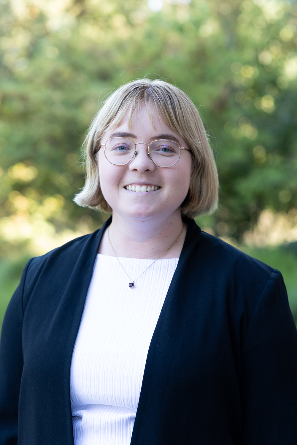 Smiling headshot of young woman with short blonde hair and glasses, black blazer, green background