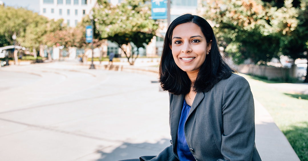 Professor Nila Bala wearing a grey suit and sitting outside in the sunshine.