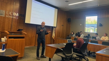 Lecturer standing in wood-paneled classroom presenting a projected slide to seated students
