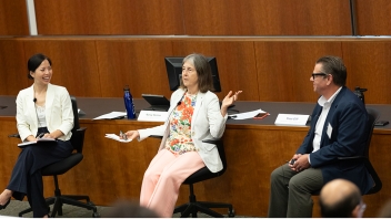 Three panelists on a wood-paneled lecture stage; center woman animatedly gestures