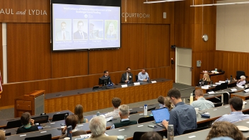 University lecture hall panel discussion with four speakers at front and seated audience.