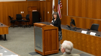 Photograph of a woman speaking at a wooden lectern in a courtroom with American flag
