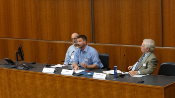 Three men seated at a wood-paneled conference table; center man gestures while speaking.