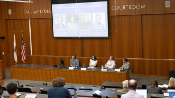 Wood-paneled courtroom with four panelists, audience, U.S. flags