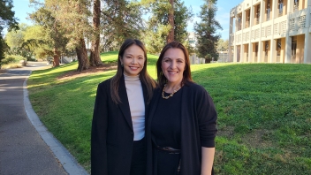 Two smiling women in dark jackets on a sunny campus walkway with trees and building.