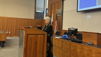 Woman speaking at wooden podium in courtroom with U.S. flag and projection screen