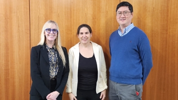 Three smiling professionals—two women and a man—standing before a wood-paneled wall.