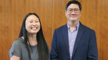 Two smiling adults, woman with long hair and man in glasses and blazer, before wood paneling