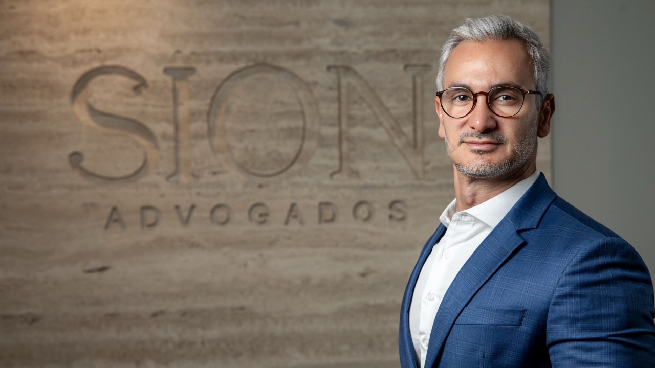 Man in blue suit and glasses by wall sign "SION ADVOGADOS".