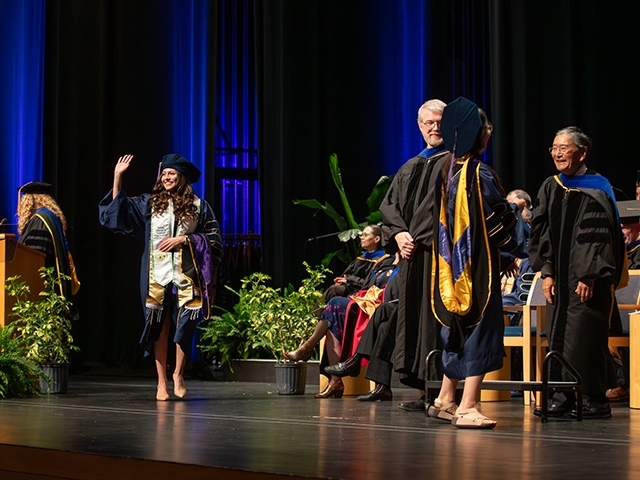 A 2025 graduate crossing the stage at the Mondavi Center during commencement and waving at the crowd. 