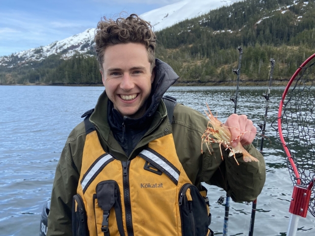 Man on a boat holds a shrimp he just caught before a background of mountains and cloudy skies. 