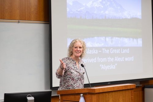A woman stands at a podium before a video screen in a classroom. 