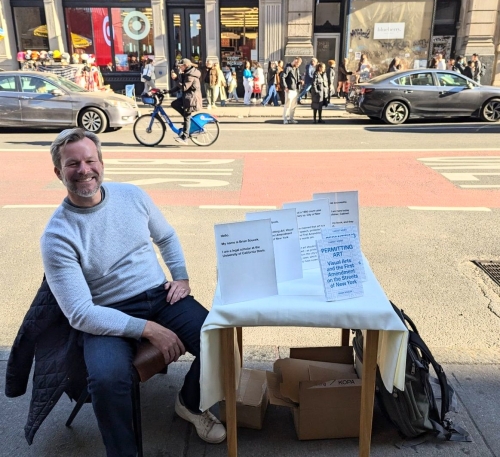 A man sits on a New York sidewalk selling books from a table in front of him.