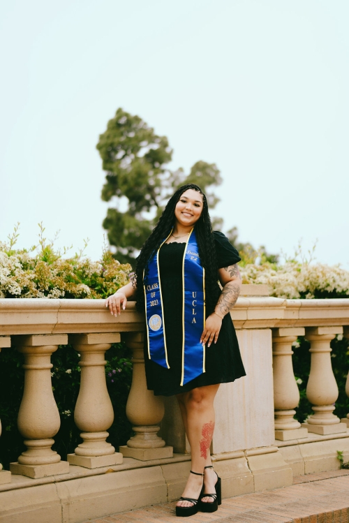 Young smiling graduate in blue stole and black dress, leaning on stone balustrade outdoors