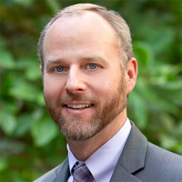 Headshot of smiling bearded man in suit against blurred green foliage
