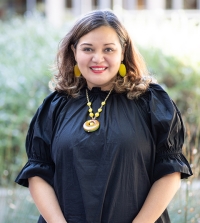 Professor Raquel Aldana wearing a black dress with a gold necklace. She is standing in the King Hall courtyard and smiling at the camera.