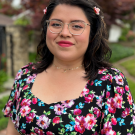 A woman wearing glasses and a colorful blouse with flowers. She is smiling at the camera.