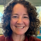 Rebekah Grodsky is smiling at the camera. She has curly dark hair, wearing a maroon sweater, in front of a blurred background.
